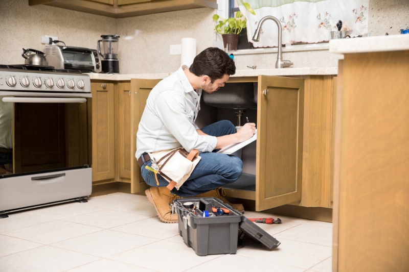 What Should I Do About Frozen Pipes? Image is a photograph of a plumbing professional looking under a kitchen sink.